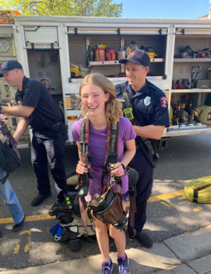 a blonde girl laughs while trying on fireman's gear while firemen stand behind her
